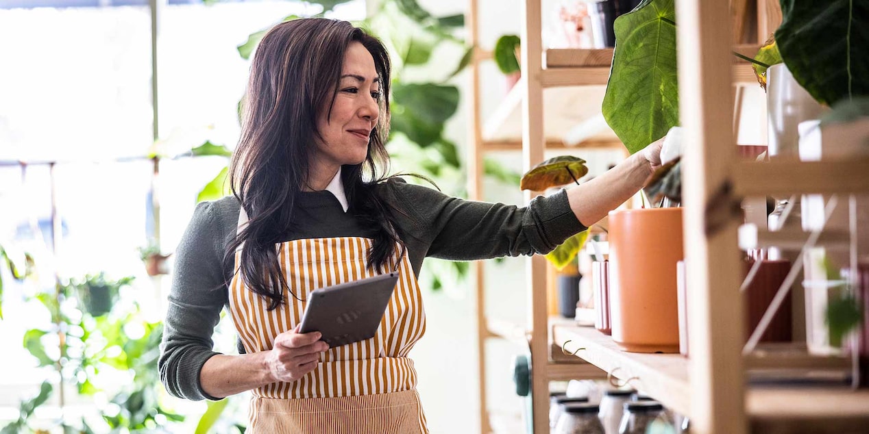 A Woman Holding a Tablet and Reaching for an Item on a Shelf