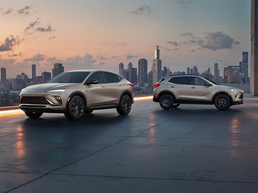 The 2026 Envista Sport Touring SUV and the 2026 Encore GX Sports Touring SUV both in the Color Ceramic Parked on a Parking Garage Rooftop Overlooking a City Skyline
